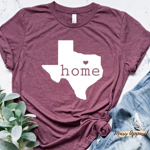 May include: A maroon t-shirt with a white outline of the state of Texas and the word "home" inside. A small pink heart is in the center of the state.