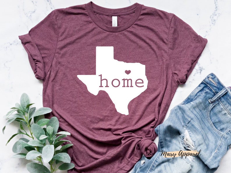 May include: A maroon t-shirt with a white outline of the state of Texas and the word "home" inside. A small pink heart is in the center of the state.