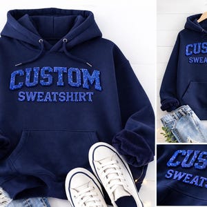 a navy blue hoodie with the text "CUSTOM SWEATSHIRT" printed on it, along with a pair of white sneakers and a pair of blue jeans.