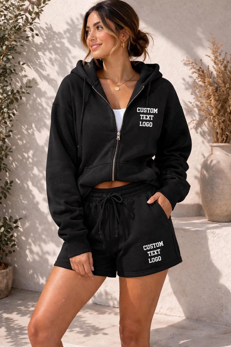 A woman wearing a black hoodie with the text "CUSTOM TEXT LOGO" and matching shorts stands in front of a white wall with a plant.