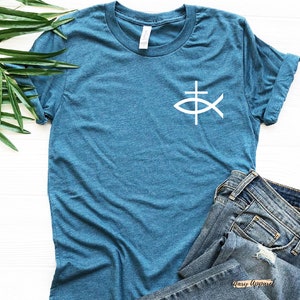 May include: A teal blue t-shirt with a white fish symbol with a cross inside. The shirt is folded and laid on a white surface with a pair of blue jeans.