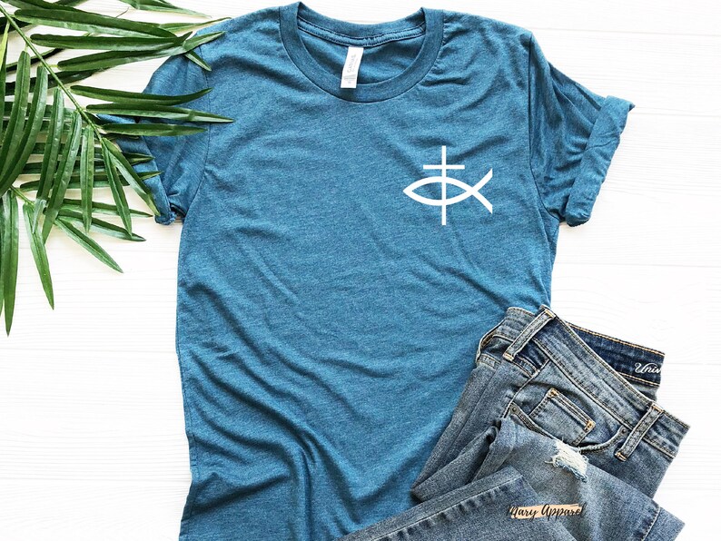 May include: A teal blue t-shirt with a white fish symbol with a cross inside. The shirt is folded and laid on a white surface with a pair of blue jeans.