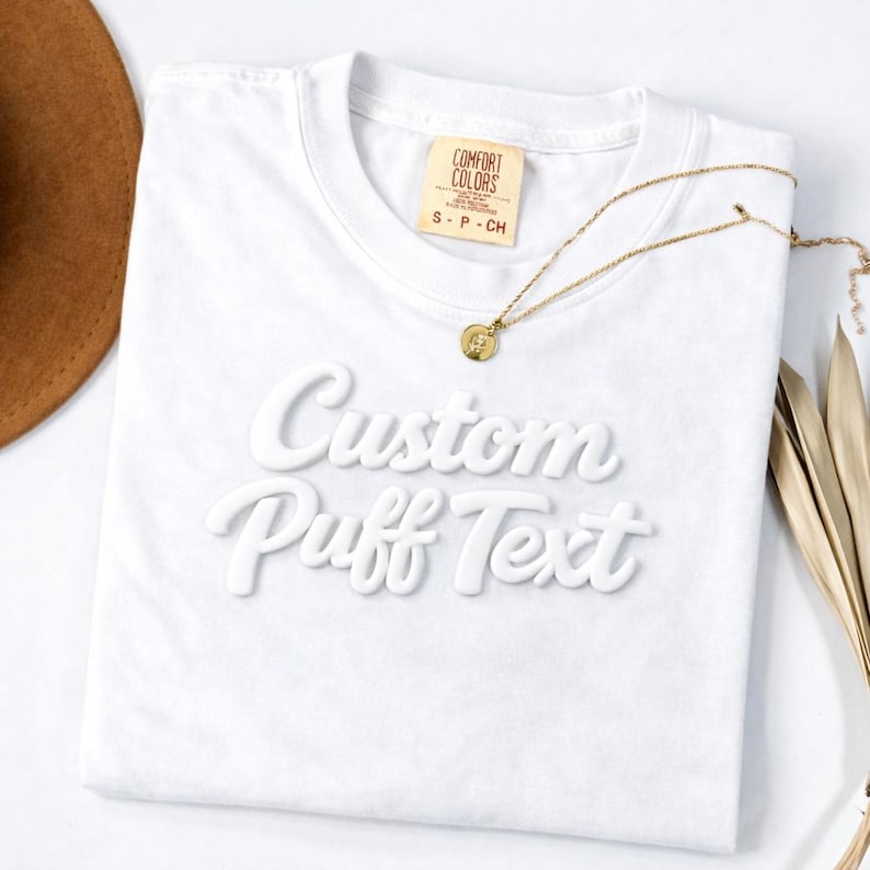 A white t-shirt with the text "Custom Puff Text" printed on it, along with a gold necklace and a brown hat.