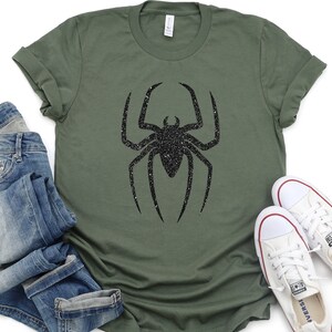 K&ouml;nnte beinhalten: Ein gr&uuml;nes T-Shirt mit einem schwarzen Glitzer-Spinnen-Motiv. Das Shirt wird mit einer blauen Jeans und wei&szlig;en Turnschuhen kombiniert.
