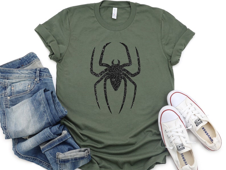 K&ouml;nnte beinhalten: Ein gr&uuml;nes T-Shirt mit einem schwarzen Glitzer-Spinnen-Motiv. Das Shirt wird mit einer blauen Jeans und wei&szlig;en Turnschuhen kombiniert.