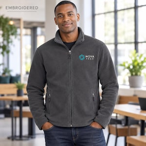 May include: A charcoal gray full-zip fleece jacket featuring a stand-up collar, long sleeves, and two zippered side pockets. The left chest displays an embroidered teal hexagon logo with the white text NOVA TECH underneath.