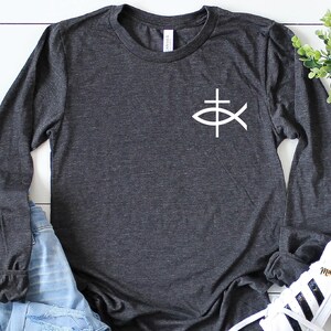 May include: A dark heather grey long-sleeve t-shirt with a white graphic of a fish with a cross on top. The shirt is laying on a white wooden surface with a pair of white sneakers and blue jeans.