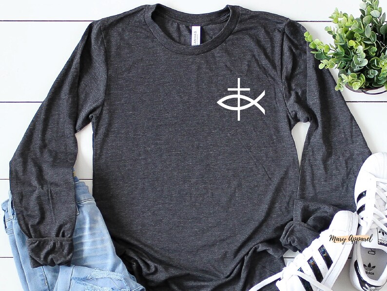 May include: A dark heather grey long-sleeve t-shirt with a white graphic of a fish with a cross on top. The shirt is laying on a white wooden surface with a pair of white sneakers and blue jeans.