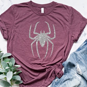 K&ouml;nnte beinhalten: Ein maronfarbenes T-Shirt mit einer silbernen Glitzer-Spinnengrafik. Das Shirt ist aus einem weichen, bequemen Material gefertigt und hat eine lockere Passform.