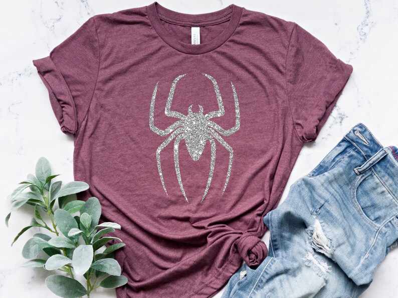 K&ouml;nnte beinhalten: Ein maronfarbenes T-Shirt mit einer silbernen Glitzer-Spinnengrafik. Das Shirt ist aus einem weichen, bequemen Material gefertigt und hat eine lockere Passform.