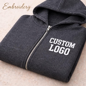 May include: A dark charcoal gray zip-up hoodie featuring a hood and a silver-toned metal zipper. The text "CUSTOM LOGO" is embroidered in white block letters on the left chest area. The word "Embroidery" is printed in the upper left corner.