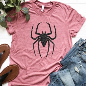 K&ouml;nnte beinhalten: Ein pinkfarbenes T-Shirt mit einem schwarzen Glitzer-Spinnen-Motiv. Das Shirt liegt auf einer wei&szlig;en Oberfl&auml;che mit einem Paar braunen Sandalen und einer blauen Jeans.