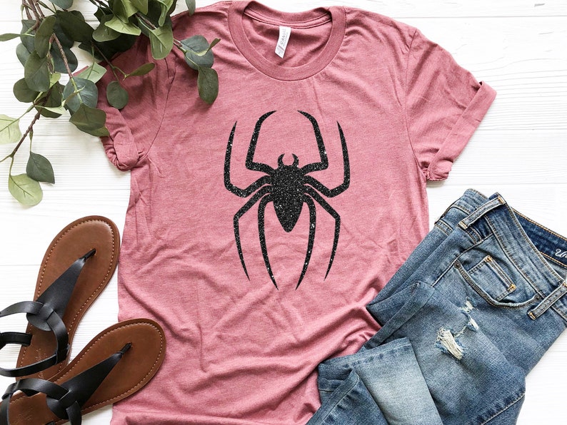 K&ouml;nnte beinhalten: Ein pinkfarbenes T-Shirt mit einem schwarzen Glitzer-Spinnen-Motiv. Das Shirt liegt auf einer wei&szlig;en Oberfl&auml;che mit einem Paar braunen Sandalen und einer blauen Jeans.