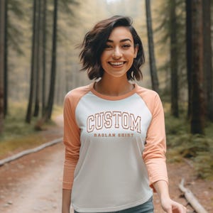 May include: A white and peach long-sleeved baseball tee with the text "CUSTOM RAGLAN SHIRT" printed on the front. The model is wearing the shirt in a woodland setting.