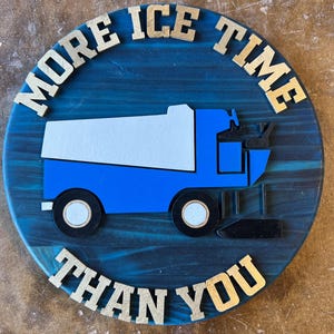May include: A round, blue wooden sign with the text "MORE ICE TIME THAN YOU" in gold letters. A blue and white ice resurfacer is in the center of the sign. The sign is on a wooden surface.