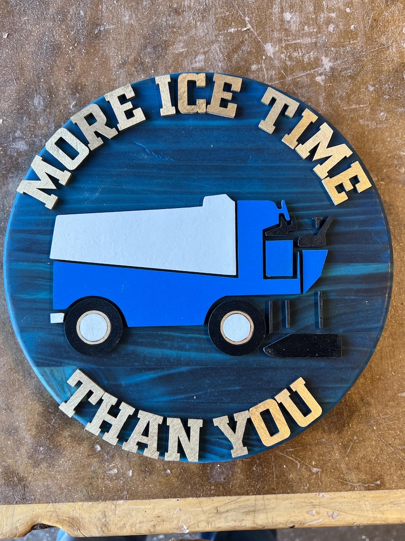 May include: A round, blue wooden sign with the text "MORE ICE TIME THAN YOU" in gold letters. A blue and white ice resurfacer is in the center of the sign. The sign is on a wooden surface.