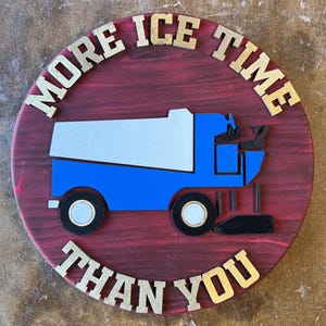 May include: A round wooden sign with a red stained background and gold lettering. The sign features a blue and white ice resurfacer graphic and the text "MORE ICE TIME THAN YOU".