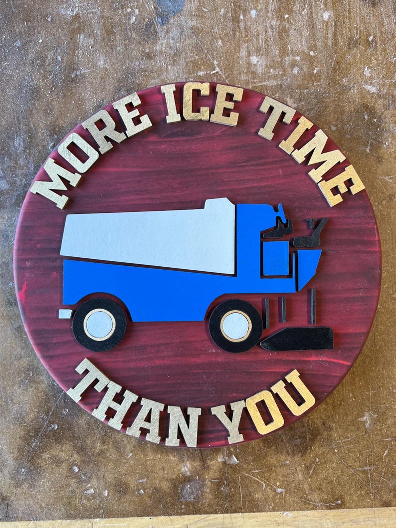 May include: A round wooden sign with a red stained background and gold lettering. The sign features a blue and white ice resurfacer graphic and the text "MORE ICE TIME THAN YOU".
