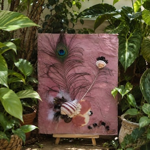 Pode incluir: Um painel retangular de textura rosa exibido num pequeno cavalete de madeira. O painel é decorado com uma grande pena de pavão, várias conchas em tons de branco, castanho e rosa, pequenas pinhas e fibras brancas dispostas centralmente.