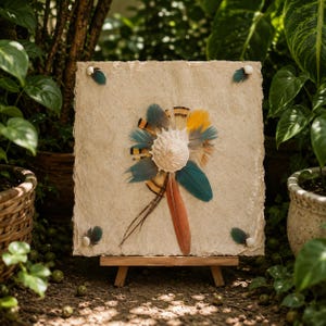 Pode incluir: Um painel quadrado de papel texturizado em tom off-white montado num pequeno cavalete de madeira. O centro apresenta uma concha branca rodeada por um arranjo radial de penas azuis, laranja, amarelas e castanhas. Quatro pequenas penas estão fixadas nos cantos.