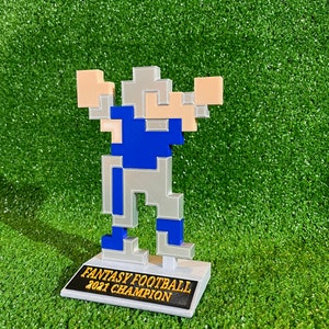 May include: A 3D printed trophy of a pixelated football player in blue and grey, standing on a white base with the text "Fantasy Football 2021 Champion" in gold lettering.