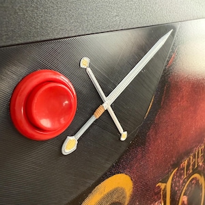 May include: A red button with a silver sword design on a black background. The sword has a brown handle and gold accents. The text "The Lord" is visible in the background.