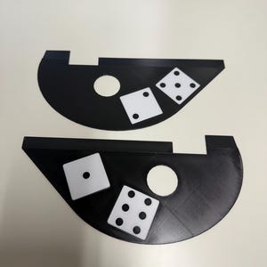 May include: Two black dice trays with white dice. Each tray has a circular hole and two dice with different dot configurations. The trays are on a white surface, with a keyboard visible in the background.