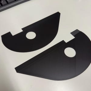 May include: Two black, semi-circular objects with a hole in the center and a rectangular tab. The objects are on a white surface, near a keyboard and mouse. The objects appear to be components or parts.