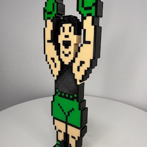 May include: A three-dimensional pixel art figure of a boxer with arms raised in victory. The figure features a black tank top, green boxing shorts, green gloves, and green boots, constructed with a blocky, layered design in black, green, and beige.