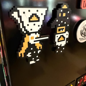 May include: Two pixelated magnets, one white and black knight with a sword, and a black and white knight. The magnets are attached to a black surface. The magnets are likely for a refrigerator or other metal surface.