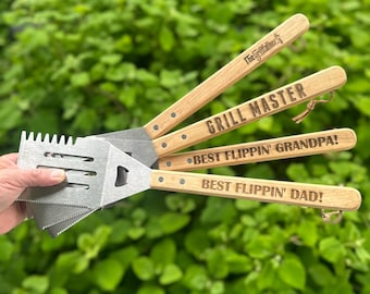 May include: A set of stainless steel grilling tools with wooden handles. The handles are engraved with the phrases "The Grillfather", "GRILL MASTER", "BEST FLIPPIN' GRANDPA!", and "BEST FLIPPIN' DAD!". The tools are displayed against a backdrop of green foliage.
