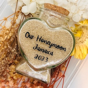 Pode incluir: Um frasco de vidro em forma de coração cheio de areia, com uma tampa de cortiça. O frasco tem um rótulo que diz "Our Honeymoon Jamaica 2023".