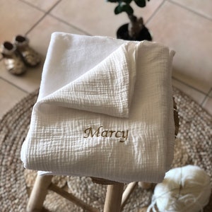 Peut inclure: Une couverture en mousseline de coton blanche avec le nom "Marcy" brodé en fil doré. La couverture est pliée et posée sur un tabouret en bois.
