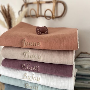 Peut inclure: Une pile de sept langes en mousseline personnalisés dans différentes couleurs, dont pêche, beige, mauve, blanc, vert clair, bleu et vert. Chaque lange porte un nom différent brodé en fil doré : Joane, Tiana, Maëli, Sajou, Basil, Noah et Eliot.