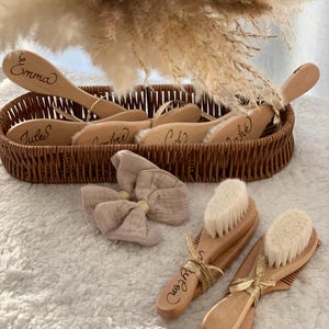 Puede incluir: Un conjunto de cepillos y peines de madera para bebés, algunos con nombres grabados, presentados en una cesta tejida. Dos juegos están fuera de la cesta, atados con cinta dorada. También hay un lazo rosa claro.