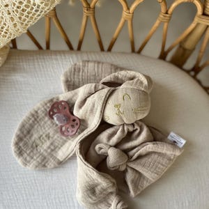 Puede incluir: Un conjunto de confort para beb&eacute; beige con una cabeza de conejo de peluche con detalles bordados y un cuerpo anudado. Incluye un babero a juego y un chupete rosa. El conjunto est&aacute; hecho de tela suave y texturizada, ideal para reci&eacute;n nacidos.