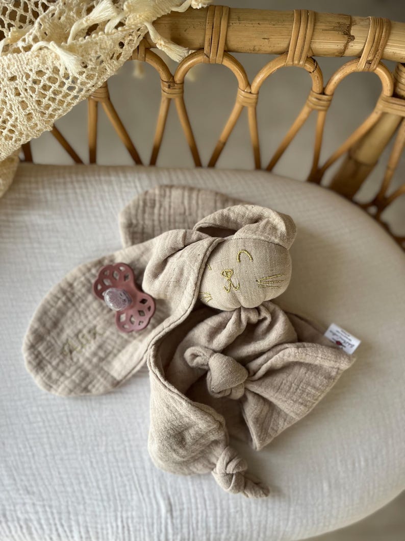 Puede incluir: Un conjunto de confort para beb&eacute; beige con una cabeza de conejo de peluche con detalles bordados y un cuerpo anudado. Incluye un babero a juego y un chupete rosa. El conjunto est&aacute; hecho de tela suave y texturizada, ideal para reci&eacute;n nacidos.