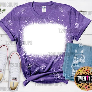 Könnte beinhalten: Lilafarbenes T-Shirt mit einem weißen Bleichmittel-Design. Das Shirt hat einen Used-Look und ist perfekt zum Personalisieren mit deinem eigenen Design. Das Shirt liegt auf einer weißen Holzoberfläche mit einem Paar weißen Sneakers und einer blauen Jeans.