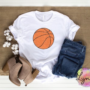 Peut inclure: T-shirt blanc avec un motif de ballon de basket-ball orange et des lignes noires, avec le texte "Basketball Mom". Le t-shirt est associ&eacute; &agrave; un jean bleu et des bottines beiges. L'image est sur une surface marron.
