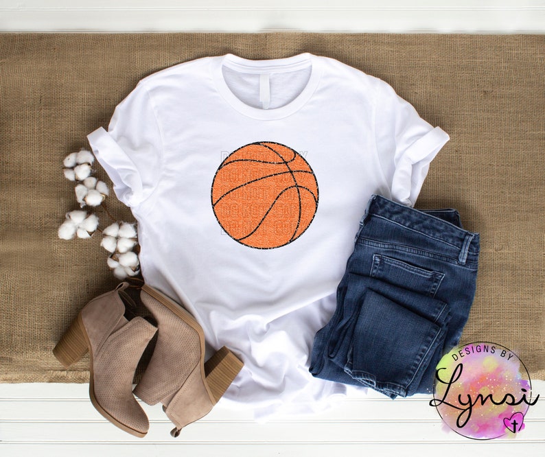 Peut inclure: T-shirt blanc avec un motif de ballon de basket-ball orange et des lignes noires, avec le texte "Basketball Mom". Le t-shirt est associ&eacute; &agrave; un jean bleu et des bottines beiges. L'image est sur une surface marron.