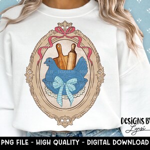 May include: White sweatshirt featuring a framed illustration of a blue hen in a basket with a rolling pin and cutting board. The frame is tan with a pink ribbon. Text at the bottom reads "PNG FILE - HIGH QUALITY - DIGITAL DOWNLOAD".