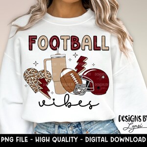 May include: White sweatshirt with the text "FOOTBALL vibes" in maroon and white. The design includes a football, a coffee cup, a leopard print heart, and a lightning bolt.