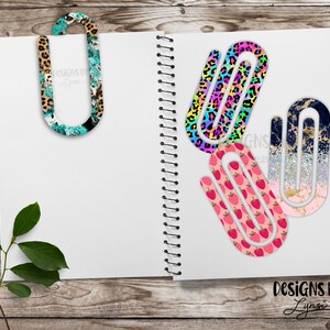 May include: A collection of decorative paper clips with various patterns, including leopard print, rainbow leopard, strawberries, and a gold and navy design. The clips are displayed on a white notebook, with the text "DESIGNS BY Lynsi".