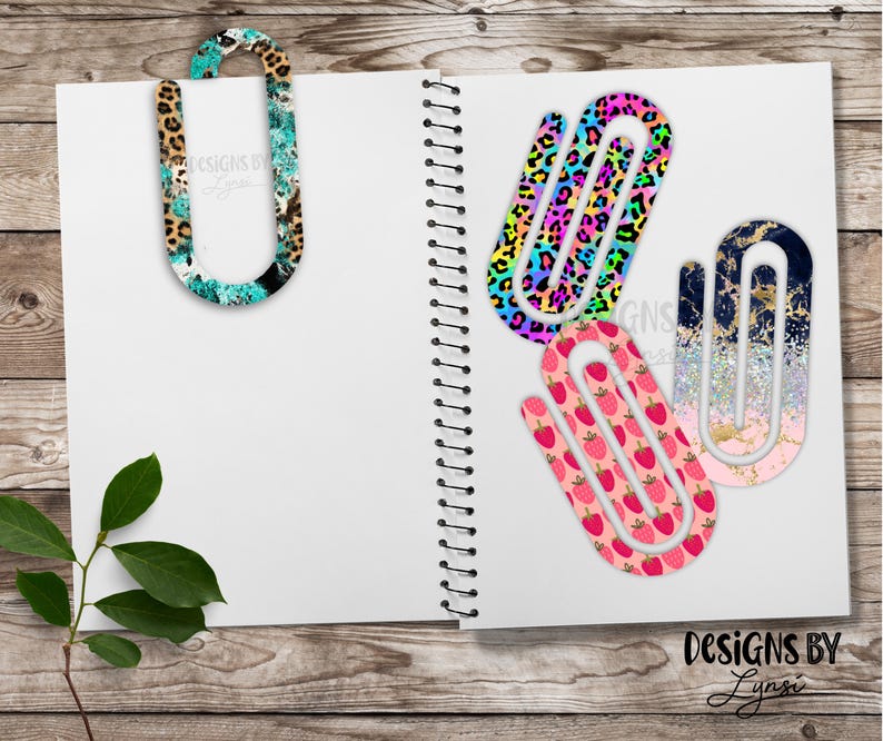 May include: A collection of decorative paper clips with various patterns, including leopard print, rainbow leopard, strawberries, and a gold and navy design. The clips are displayed on a white notebook, with the text "DESIGNS BY Lynsi".
