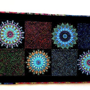 May include: A black table runner with a repeating pattern of colourful, stylised floral designs and speckled squares. The runner's edges are trimmed with a patterned border. The design incorporates shades of blue, teal, red, and green.