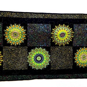 May include: A black quilted table runner featuring a repeating pattern of colorful, stylized floral designs. The runner has a black border with a dotted pattern and a series of square and circular designs in shades of yellow, green, and blue.