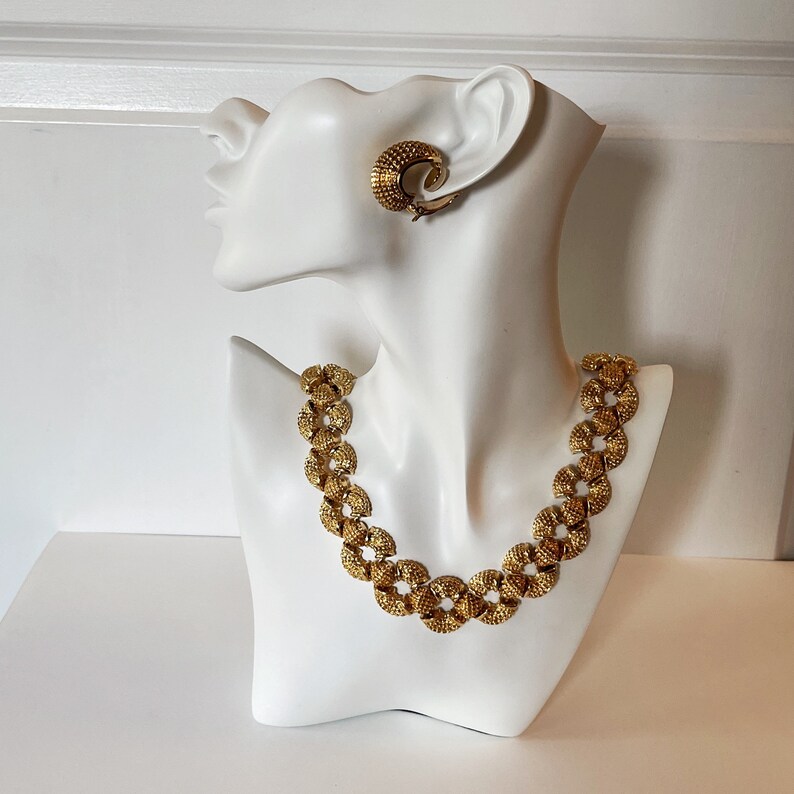 May include: A gold-tone necklace and earrings set with a textured, circular design. The necklace is made of linked circles and the earrings are hoop-shaped.