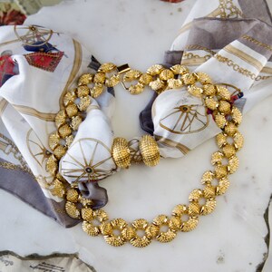 May include: A gold necklace with round, textured links. The necklace is displayed on a white marble surface with a patterned scarf.