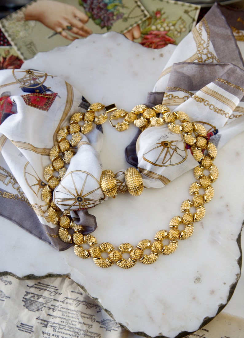 May include: A gold necklace with round, textured links. The necklace is displayed on a white marble surface with a patterned scarf.
