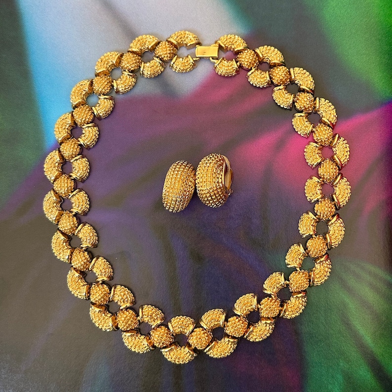May include: A gold necklace and earrings set with a textured, circular design. The necklace is made of linked circles and the earrings are hoop-shaped.